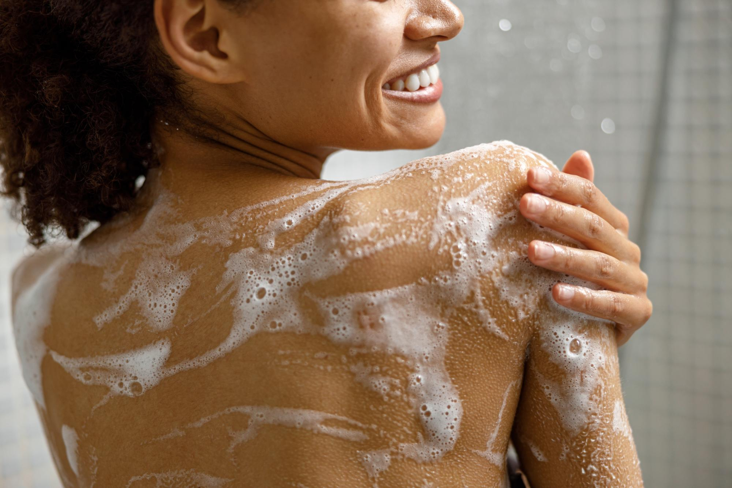 Gros plan sur une femme souriante prenant une douche avec du gel ou de la mousse de shampoing dans la salle de bain
